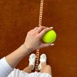 Ръка с тенис топка на тенис корт, носеща стилна гривна на фона на оранжево покритие.