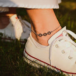 Модерна гривна за глезен от медицинска стомана с черни перли, носена с бели маратонки на зелена трева.