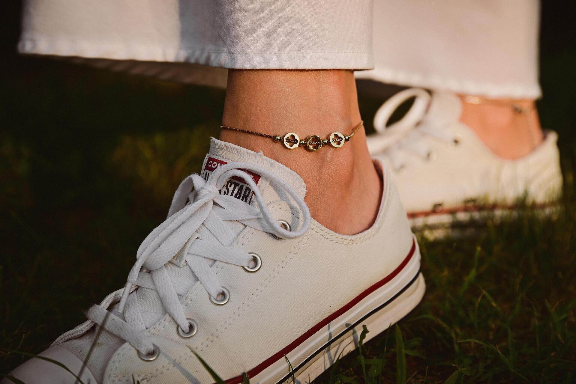 Гривна за глезен от медицинска стомана в златист цвят, носена с бели маратонки на фон от зелена трева.